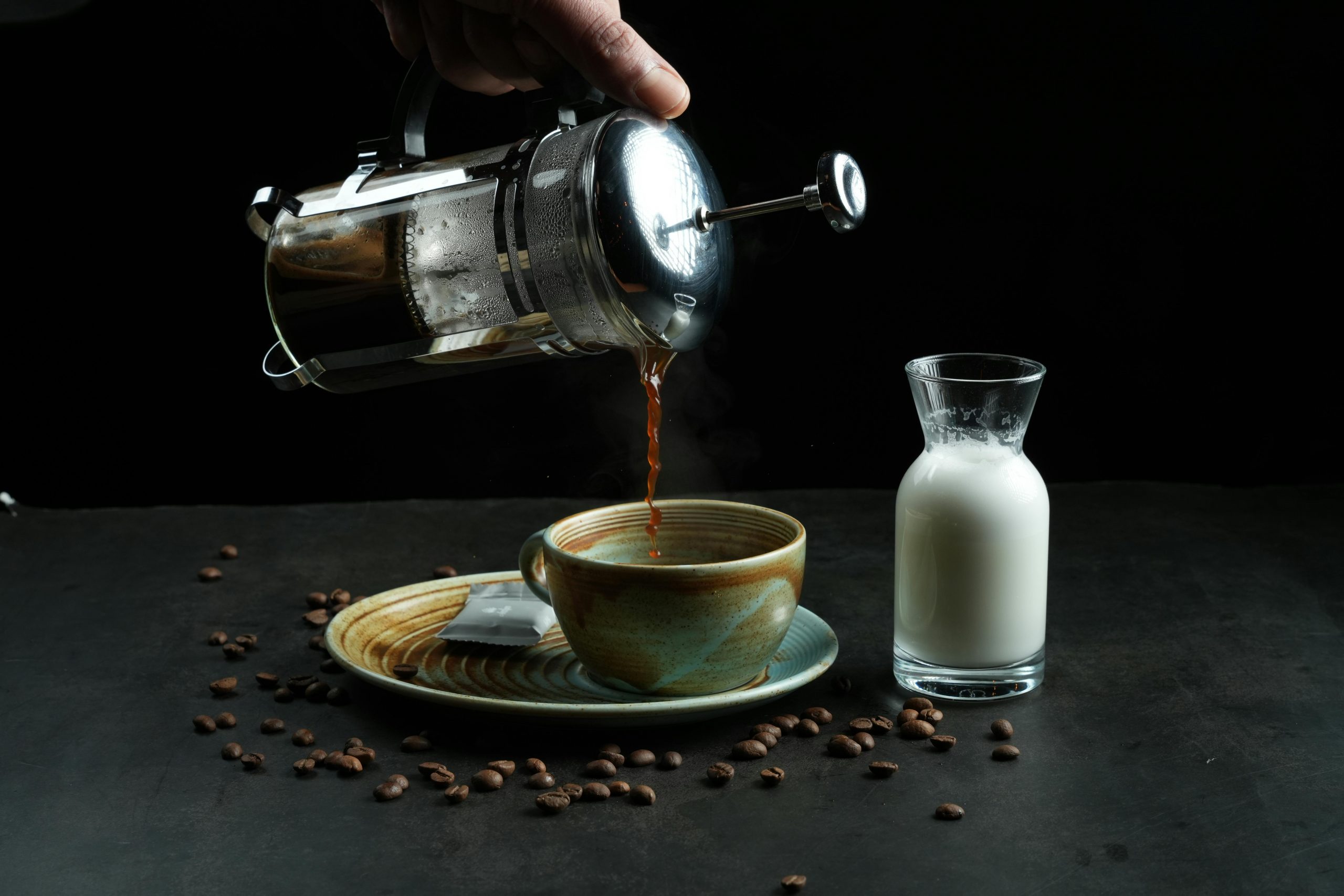 french press coffee being poured into a cup on a saucer, with milk and coffee beans next to it